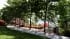 CGI of a tree-shaded gravel terrace with a bistro table, red flowering shrubs and a palm framing the view of Lake Como